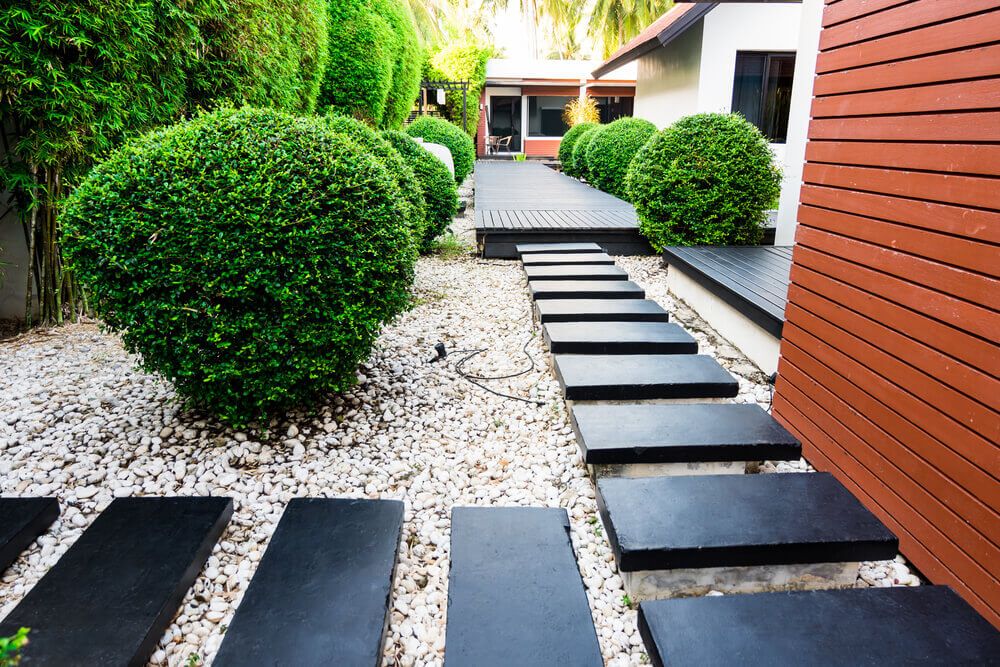 Black stones garden path on white pebbles and lush green trees.